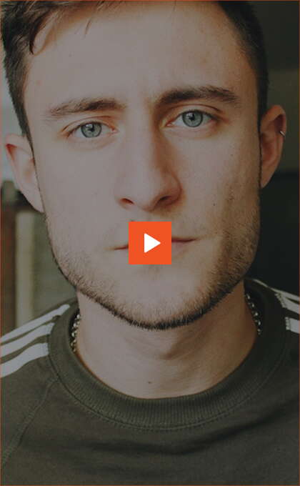 Close-up of a young man with blue eyes and short brown hair, wearing a dark green shirt with white stripes on the shoulders. An orange play button icon is centered on the image.
