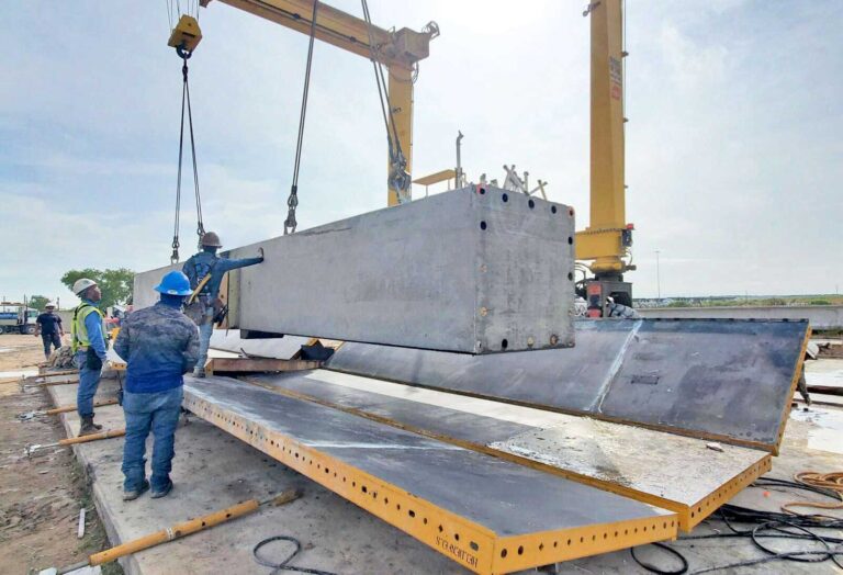 Construction workers guide a large concrete beam being lifted by a crane onto a steel structure at a construction site. Safety gear like helmets and vests are worn, and tools and cables are visible on the ground.